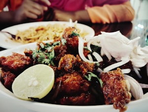 A delicious plate of spicy fried meat garnished with fresh cilantro, sliced onions, and a wedge of lime. The background shows a dish of noodles or rice and two people, one wearing red and the other orange, with their hands visible.