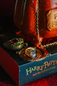 A time-turner necklace with intricate details and a locket featuring a stylized skull motif are placed on a book titled 'Harry Potter and the Goblet of Fire.' The background features a large, glossy, orange ceramic object with a visible plaque and chain. The setting suggests a magical or mystical theme.