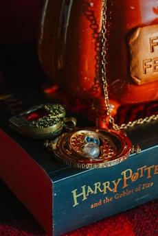 A time-turner necklace with intricate details and a locket featuring a stylized skull motif are placed on a book titled 'Harry Potter and the Goblet of Fire.' The background features a large, glossy, orange ceramic object with a visible plaque and chain. The setting suggests a magical or mystical theme.