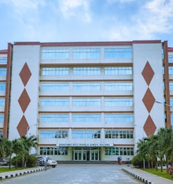 A multi-story building features a symmetrical design with two prominent diamond-shaped designs on its façade. The structure has several rows of large windows, and at the entrance, there is a sign labeled 'Babatunde Raji Fashola Senate Building'. Several palm trees line the path leading to the entrance, and a few people are seen near the doorway.