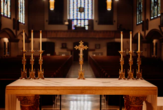 A church altar with a central ornate cross flanked by two sets of three tall white candles in elaborate gold holders. The background features rows of empty pews and large stained-glass windows, creating a serene and solemn atmosphere.