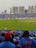 A packed cricket stadium with large crowds wearing blue jerseys, focused on a cricket match taking place on the field. High-rise buildings can be seen in the background, and banners are displayed around the stadium. Spectators are enthusiastic, capturing moments with their phones.