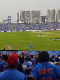 A packed cricket stadium with large crowds wearing blue jerseys, focused on a cricket match taking place on the field. High-rise buildings can be seen in the background, and banners are displayed around the stadium. Spectators are enthusiastic, capturing moments with their phones.