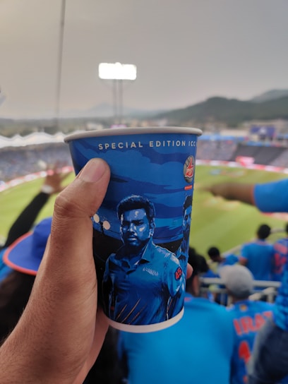 A person holding a blue cup with a special edition ICC design featuring a cricket player. The background shows a sports stadium filled with spectators wearing blue jerseys, indicating the scene is likely a cricket match.