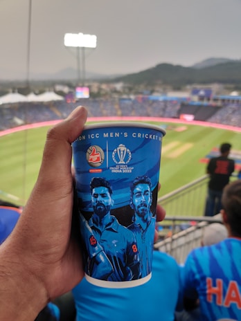 A person's hand holds a cricket-themed beverage cup featuring illustrations of two cricketers with numbers 45 and 8. The background displays a large stadium with a cricket pitch and blurred spectators under a cloudy sky.