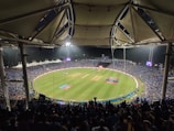 A large stadium filled with spectators watching a cricket match under bright floodlights. The field is a lush green with clearly marked pitches and boundary lines. The stadium has a modern architectural design with a white, tent-like roof structure. Players can be seen on the field, and the crowd creates a vibrant atmosphere.