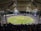 A large stadium filled with spectators watching a cricket match under bright floodlights. The field is a lush green with clearly marked pitches and boundary lines. The stadium has a modern architectural design with a white, tent-like roof structure. Players can be seen on the field, and the crowd creates a vibrant atmosphere.