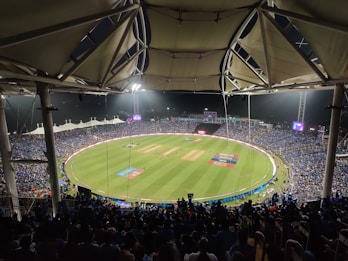 A large stadium filled with spectators watching a cricket match under bright floodlights. The field is a lush green with clearly marked pitches and boundary lines. The stadium has a modern architectural design with a white, tent-like roof structure. Players can be seen on the field, and the crowd creates a vibrant atmosphere.