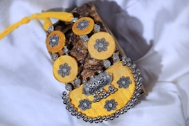 A piece of jewelry composed of circular yellow fabric elements adorned with intricate silver floral designs. The centerpiece features a larger yellow circular piece with a silver floral design, encircled with additional decorative elements. The background includes a bark-like textured item and white fabric.