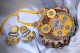 A set of intricately designed jewelry consisting of a necklace and matching earrings, with a vibrant yellow fabric backdrop. The pieces feature ornate silver floral patterns and decorative beads, presented elegantly on a wooden surface.