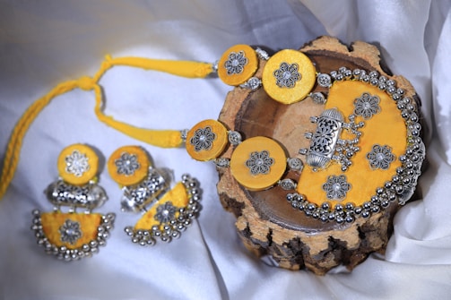 A set of intricately designed jewelry consisting of a necklace and matching earrings, with a vibrant yellow fabric backdrop. The pieces feature ornate silver floral patterns and decorative beads, presented elegantly on a wooden surface.