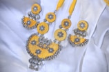 A beautifully crafted necklace featuring intricate silver floral designs set against vibrant yellow fabric. The necklace combines circular and semi-circular elements adorned with detailed silver balls and motifs, giving it a traditional and ornate appearance. It is laid out on a white fabric background, enhancing the contrast and highlighting its craftsmanship.