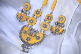 A beautifully crafted necklace featuring intricate silver floral designs set against vibrant yellow fabric. The necklace combines circular and semi-circular elements adorned with detailed silver balls and motifs, giving it a traditional and ornate appearance. It is laid out on a white fabric background, enhancing the contrast and highlighting its craftsmanship.