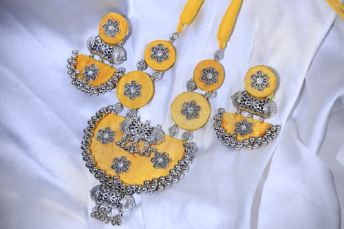 A beautifully crafted necklace featuring intricate silver floral designs set against vibrant yellow fabric. The necklace combines circular and semi-circular elements adorned with detailed silver balls and motifs, giving it a traditional and ornate appearance. It is laid out on a white fabric background, enhancing the contrast and highlighting its craftsmanship.