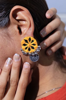 A close-up of an ear featuring a large, colorful earring. The earring has a bright yellow and black design resembling a flower, with small metallic beads and a dangling coin charm. A hand gently touches the ear, showcasing neatly manicured fingers with natural nails.