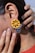 A close-up of an ear featuring a large, colorful earring. The earring has a bright yellow and black design resembling a flower, with small metallic beads and a dangling coin charm. A hand gently touches the ear, showcasing neatly manicured fingers with natural nails.