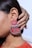 A close-up of a person's ear adorned with a large, decorative earring. The earring features intricate silver designs with floral motifs and a mix of red fabric and silver elements. A hand gently touches the earring, and the background is a soft, neutral color.