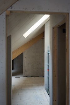 A partially built or renovated interior space with visible wooden beams and slanted ceilings. The flooring and walls appear unfinished, and there is a skylight allowing natural light to enter the room. There are also some construction materials and scaffolding visible in the image.
