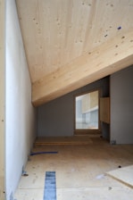 The image depicts a partially built room with an angled, unfinished wooden ceiling and walls. The floor is also wooden, with some construction materials, such as metal brackets and blue tubes, visible. A small rectangular opening is present on one wall, allowing light to filter through.