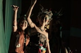 Two women are dressed in elaborate traditional costumes, adorned with intricate beadwork and headpieces featuring feathers. They appear to be engaged in a dance or performance against a dimly lit background.