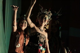 Two women are dressed in elaborate traditional costumes, adorned with intricate beadwork and headpieces featuring feathers. They appear to be engaged in a dance or performance against a dimly lit background.