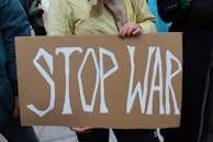 A person holding a cardboard sign with the words 'STOP WAR' written in bold white letters. The individual's hands are visible, and they are wearing a yellow jacket.