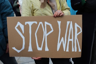 A person holding a cardboard sign with the words 'STOP WAR' written in bold white letters. The individual's hands are visible, and they are wearing a yellow jacket.
