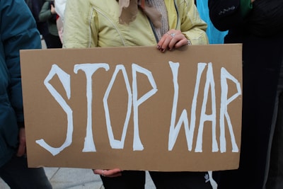 A person holding a cardboard sign with the words 'STOP WAR' written in bold white letters. The individual's hands are visible, and they are wearing a yellow jacket.