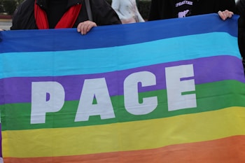 A large rainbow-colored flag features the word 'PACE' in bold white letters. Multiple horizontal stripes in vibrant colors include purple, blue, turquoise, green, yellow, and orange. Two people are partially visible, holding the top corners of the flag.