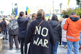 The image depicts a group of people participating in a public gathering or protest. A prominent figure in the center is wearing a dark jacket with the words 'NO WAR' written in large, bold letters on the back. Several individuals are holding flags and taking photos, suggesting it might be a demonstration.