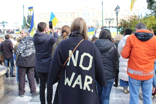 The image depicts a group of people participating in a public gathering or protest. A prominent figure in the center is wearing a dark jacket with the words 'NO WAR' written in large, bold letters on the back. Several individuals are holding flags and taking photos, suggesting it might be a demonstration.