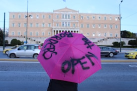 A large building with classical architecture in the background, resembling a government or historic site, with cars passing by on the street. In the foreground, a person holds a bright pink umbrella with black spray-painted text that reads 'OPEN the BORDERS.'