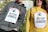 Two individuals are wearing placards with the message 'STOP RACISM' printed on them, accompanied by a handprint graphic. The person on the left is wearing a gray hoodie, while the person on the right is dressed in a yellow hoodie. The placards are attached with red tape. Graffiti and trees are visible in the background.