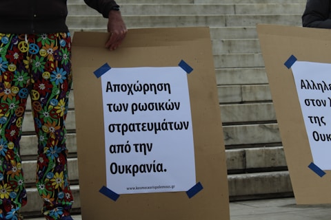 A person stands outdoors holding a large cardboard sign with Greek text advocating for the withdrawal of Russian troops from Ukraine. The person is wearing colorful pants featuring peace symbols and smiley faces. Next to them, another similar sign is partially visible. The background shows stone steps.