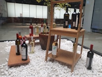 An indoor display featuring a variety of wine bottles arranged on wooden shelves and platforms. The bottles are placed on a bed of white decorative stones, with some positioned individually and others grouped together. The background includes a window with a frosted pane, revealing a tree with green leaves partially visible behind it.