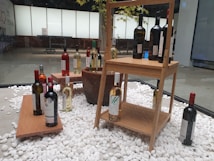 An indoor display featuring a variety of wine bottles arranged on wooden shelves and platforms. The bottles are placed on a bed of white decorative stones, with some positioned individually and others grouped together. The background includes a window with a frosted pane, revealing a tree with green leaves partially visible behind it.