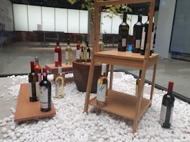 An indoor display featuring a variety of wine bottles arranged on wooden shelves and platforms. The bottles are placed on a bed of white decorative stones, with some positioned individually and others grouped together. The background includes a window with a frosted pane, revealing a tree with green leaves partially visible behind it.