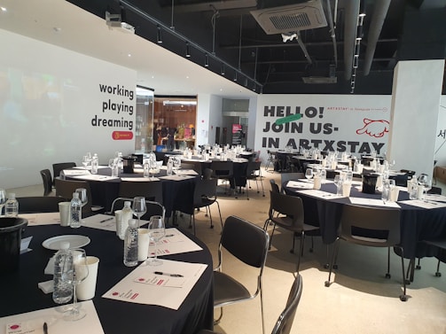 A modern, well-lit conference room is prepared for an event. Several round tables are arranged with black tablecloths, each set with water bottles, glasses, paper, and pens. The walls are adorned with motivational phrases like 'working playing dreaming' and an invitation to 'join us in ARTXSTAY.' Ceiling lights are visible, illuminating the space, and the atmosphere suggests a professional yet creative gathering.