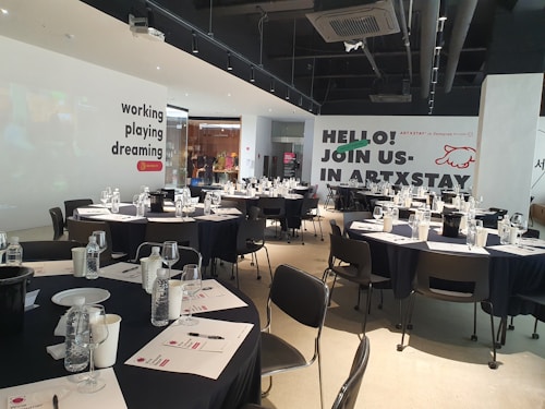 A modern, well-lit conference room is prepared for an event. Several round tables are arranged with black tablecloths, each set with water bottles, glasses, paper, and pens. The walls are adorned with motivational phrases like 'working playing dreaming' and an invitation to 'join us in ARTXSTAY.' Ceiling lights are visible, illuminating the space, and the atmosphere suggests a professional yet creative gathering.