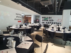 A modern, well-lit conference room is prepared for an event. Several round tables are arranged with black tablecloths, each set with water bottles, glasses, paper, and pens. The walls are adorned with motivational phrases like 'working playing dreaming' and an invitation to 'join us in ARTXSTAY.' Ceiling lights are visible, illuminating the space, and the atmosphere suggests a professional yet creative gathering.