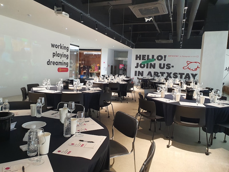 A modern, well-lit conference room is prepared for an event. Several round tables are arranged with black tablecloths, each set with water bottles, glasses, paper, and pens. The walls are adorned with motivational phrases like 'working playing dreaming' and an invitation to 'join us in ARTXSTAY.' Ceiling lights are visible, illuminating the space, and the atmosphere suggests a professional yet creative gathering.