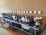 A number of wine bottles organized in a row on a long table against a beige wall. Each bottle is numbered from 1 to 16 with circular number signs placed above them. Some of the bottles are placed in ice buckets. There are multiple corks scattered across the table, and a large metal bowl containing white wine bottles is visible at one end of the table. Under the table are several baskets and another bowl.