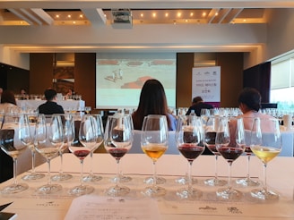 A room filled with rows of wine glasses, each containing different types of wine, set on a table with a printed guide. People are seated at tables facing a large presentation screen at the front, which displays a slide related to a wine tasting event. The atmosphere suggests a formal or organized tasting session with a focus on wine education or appreciation.