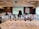 A room filled with rows of wine glasses, each containing different types of wine, set on a table with a printed guide. People are seated at tables facing a large presentation screen at the front, which displays a slide related to a wine tasting event. The atmosphere suggests a formal or organized tasting session with a focus on wine education or appreciation.