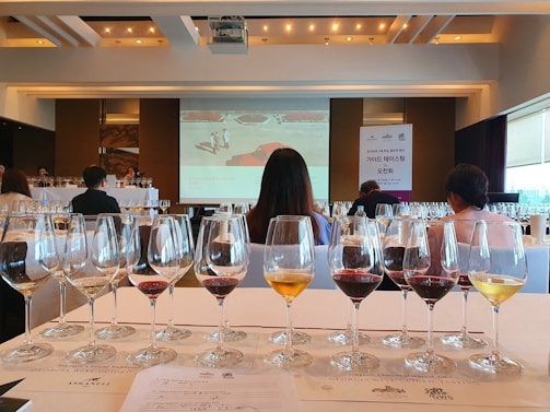 A room filled with rows of wine glasses, each containing different types of wine, set on a table with a printed guide. People are seated at tables facing a large presentation screen at the front, which displays a slide related to a wine tasting event. The atmosphere suggests a formal or organized tasting session with a focus on wine education or appreciation.