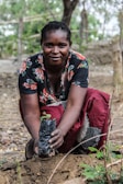 A person crouches outdoors, planting a small sapling in a plastic bag into the soil. The individual is wearing a floral patterned top and a red skirt, with a focused and content expression. The background consists of trees and natural surroundings.