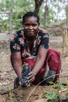 A person crouches outdoors, planting a small sapling in a plastic bag into the soil. The individual is wearing a floral patterned top and a red skirt, with a focused and content expression. The background consists of trees and natural surroundings.