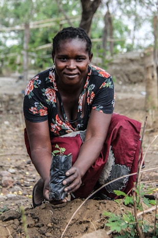 A person crouches outdoors, planting a small sapling in a plastic bag into the soil. The individual is wearing a floral patterned top and a red skirt, with a focused and content expression. The background consists of trees and natural surroundings.