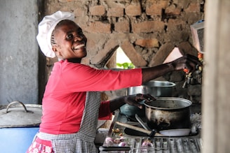 A person wearing a chef hat and a red shirt smiles while working in a rustic kitchen with stone walls. Various cooking utensils, pots, and a knife are placed on the counter next to them.
