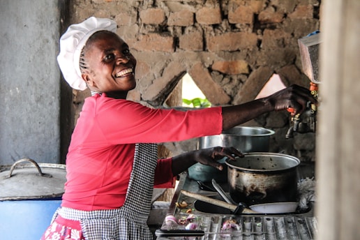 A person wearing a chef hat and a red shirt smiles while working in a rustic kitchen with stone walls. Various cooking utensils, pots, and a knife are placed on the counter next to them.